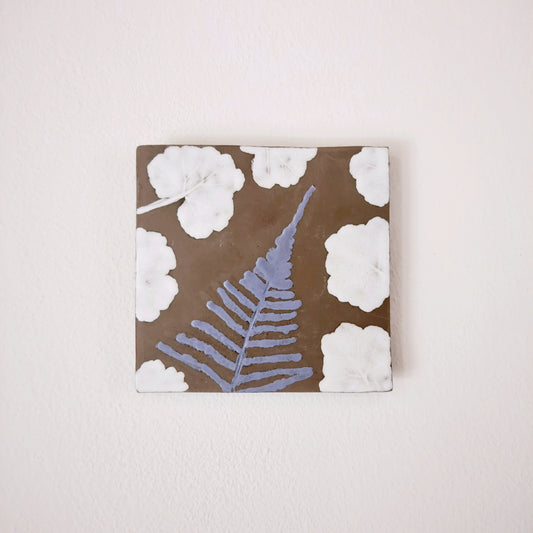 Formella in ceramica con decorazione a foglie azzurre e bianche su fondo marrone.